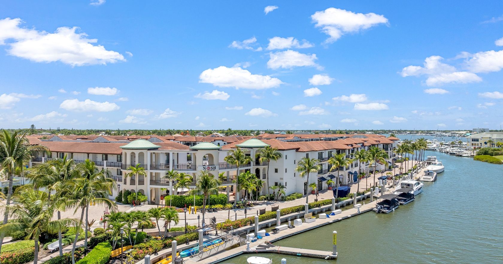 A sunny coastal marina with palm trees, white buildings, and boats docked along a canal by a blue sky&mdash;perfect day by the water.