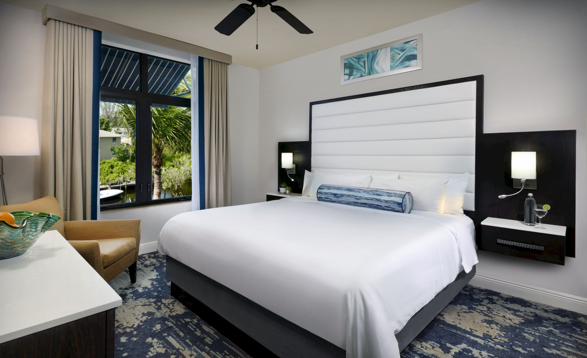 A modern hotel room with a large bed, two nightstands, a white and blue decor, a seating area, and a window with curtains.