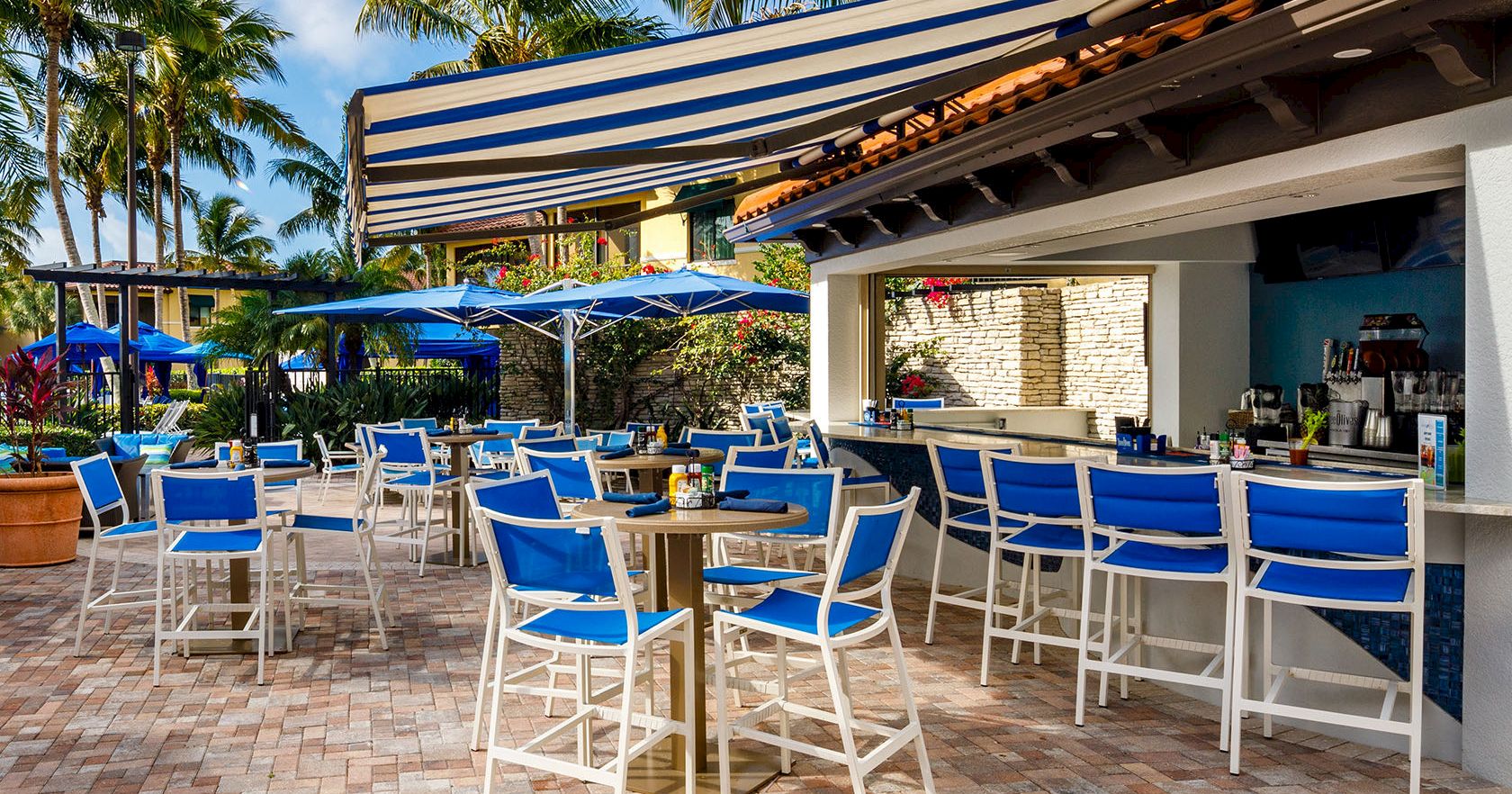 A sunny outdoor bar and patio with blue chairs, high tables, umbrellas, and a striped awning, perfect for tropical drinks and lounging.