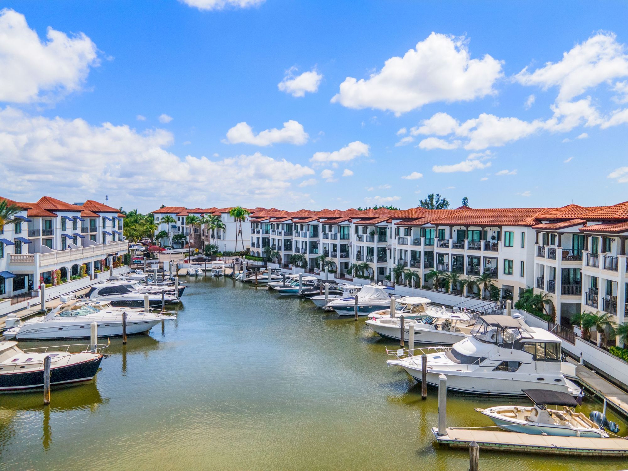 Luxurious marina with rows of white and blue townhomes, boats docked along calm water, under a bright blue sky with fluffy clouds, sunny day.