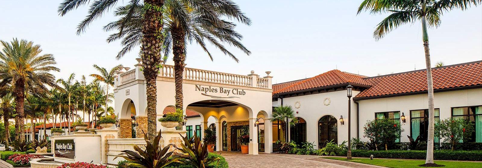 A sunny resort entrance with palm trees, a Mediterranean-style building labeled &ldquo;Naples Bay Club,&rdquo; and a landscaped, curved brick driveway.