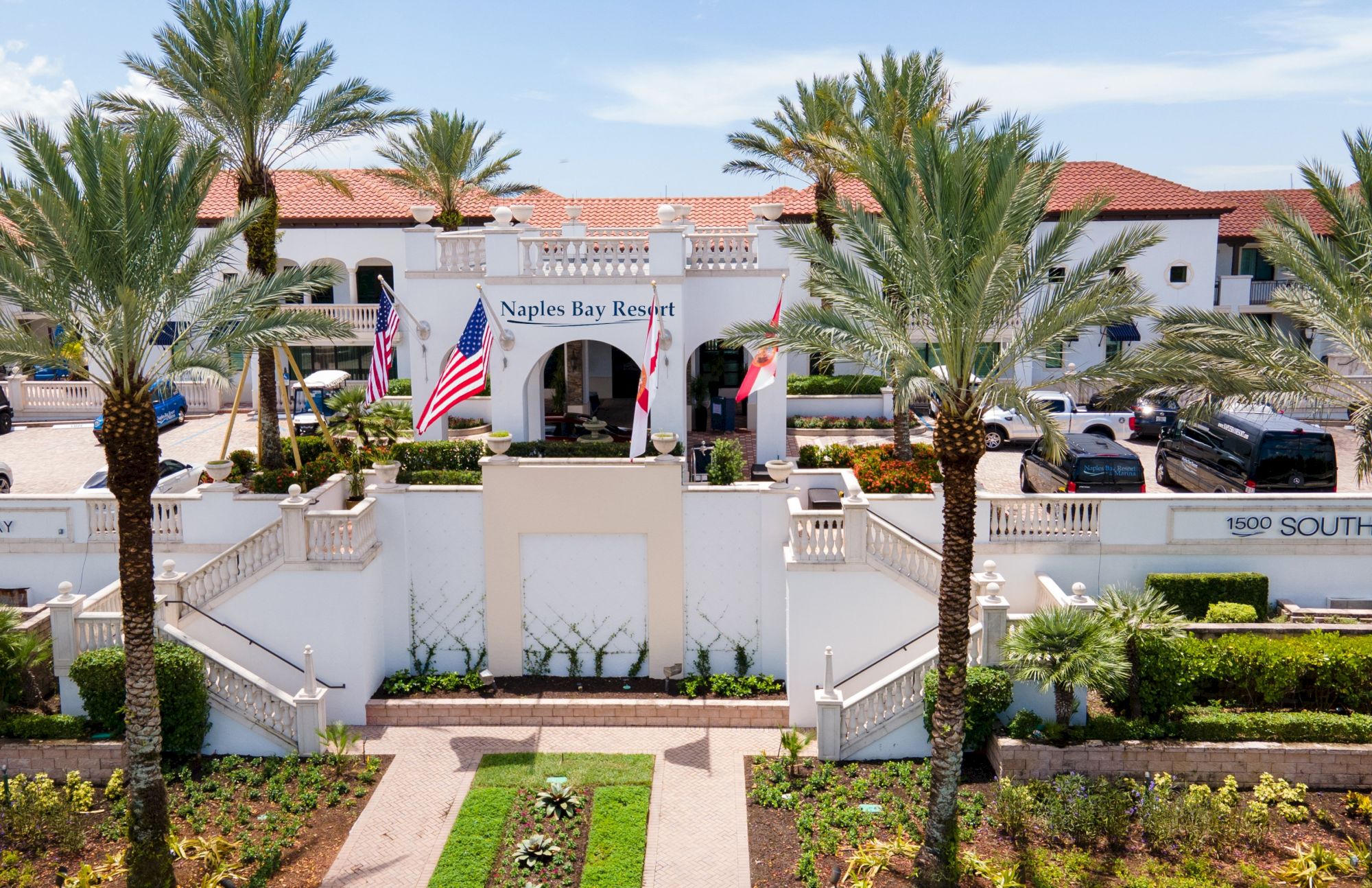 A resort-style entrance with white buildings, palm trees, and American flags at Naples Bay Resort, sunny and inviting.