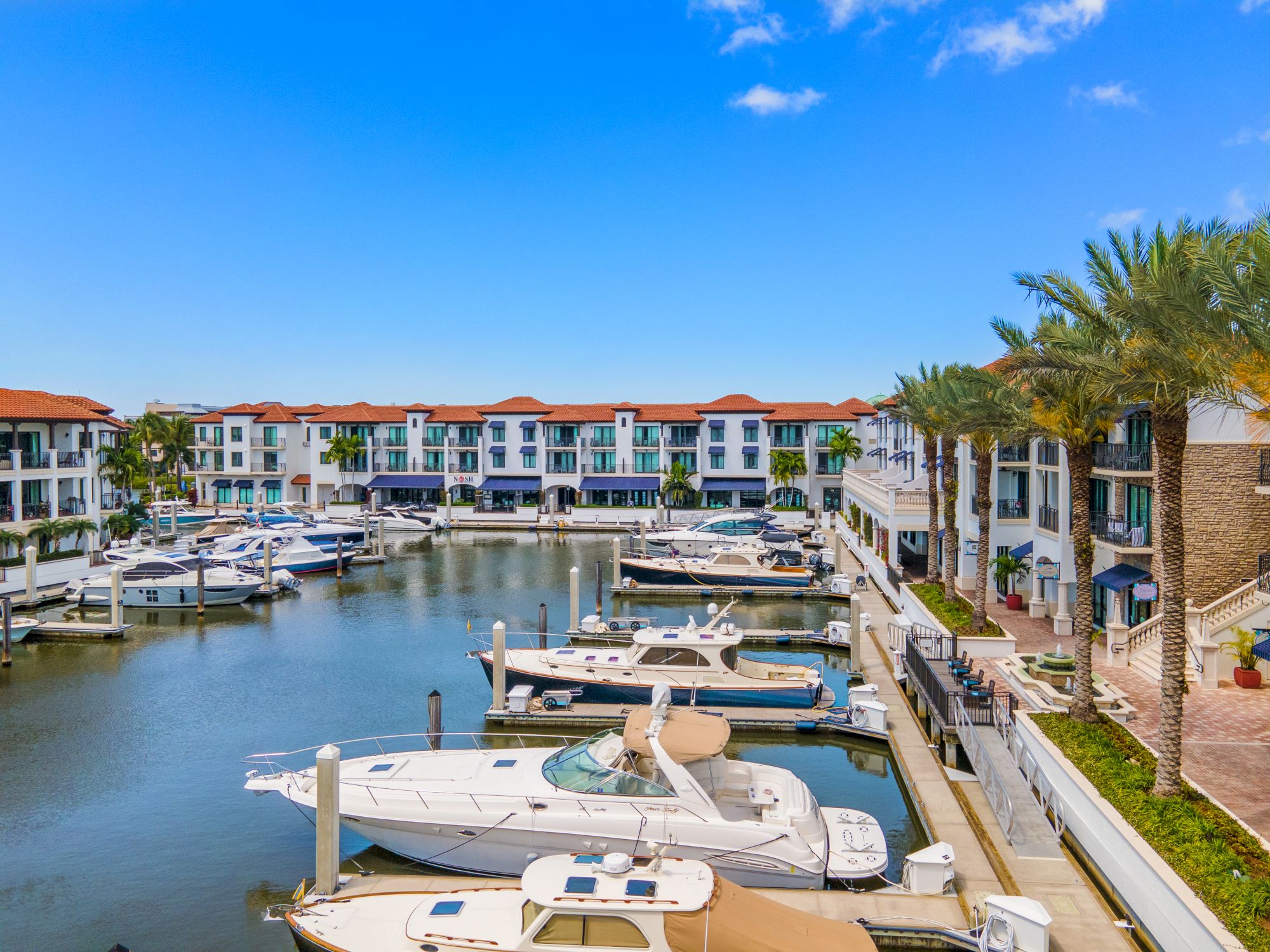 A marina with several private boats docked along calm water, surrounded by a row of colorful, low-rise residential buildings and palm trees, sunny sky.