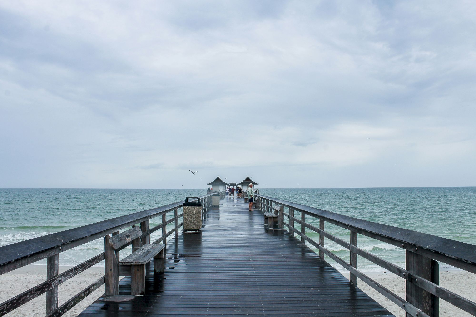 A long wooden pier extends into a calm blue sea under a cloudy sky, with small huts at the far end and railings on both sides.