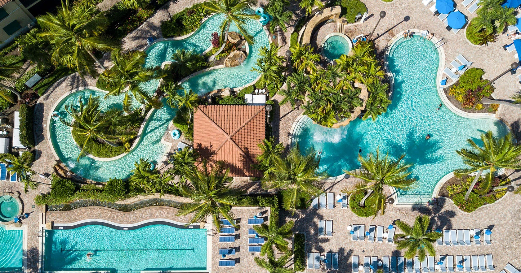 An aerial view of a resort pool area with winding pools, palm trees, sun loungers, and a central building, all surrounded by lush greenery.