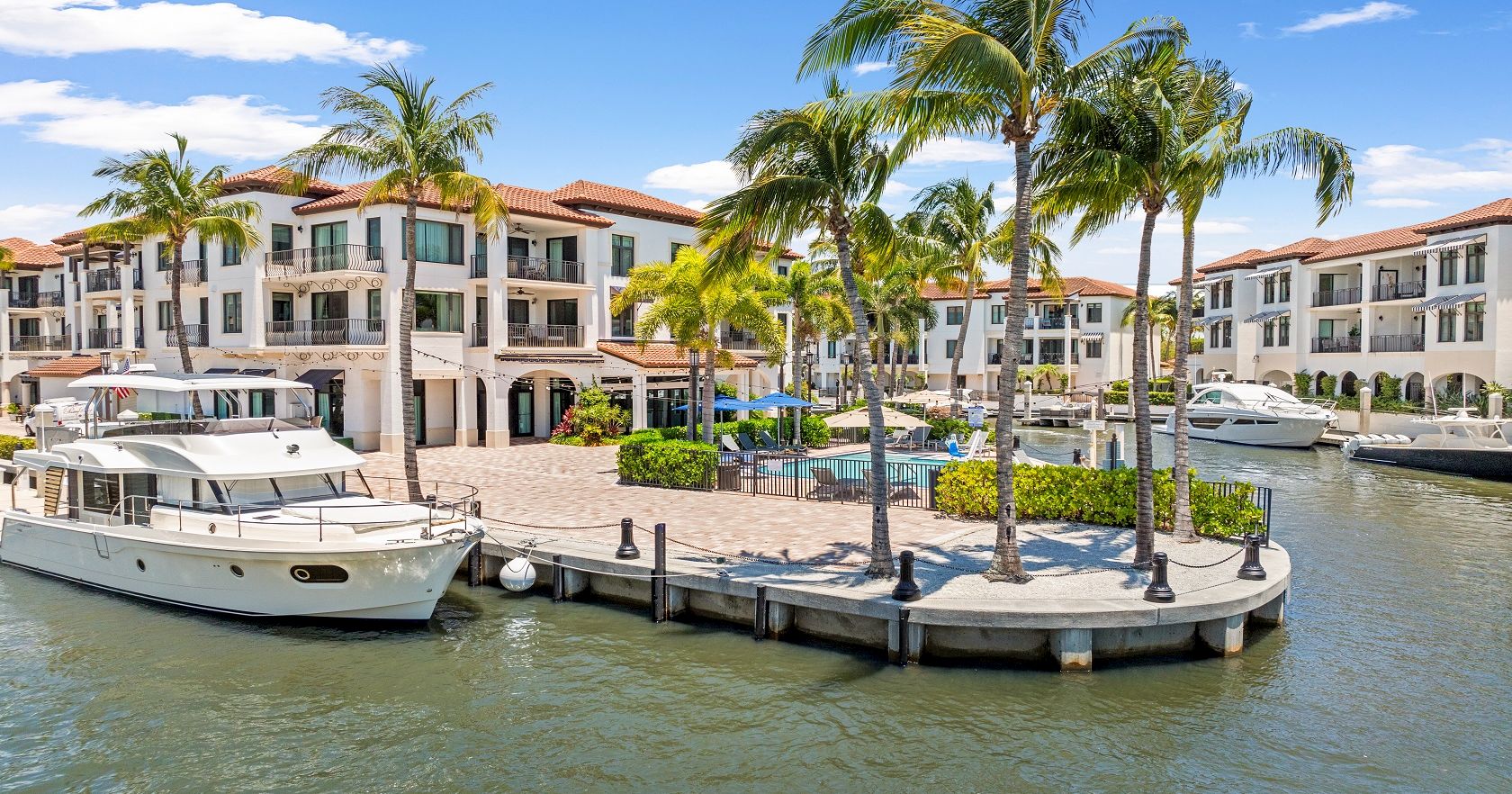 A marina with a small curved boardwalk, palm trees, boats docked, and a bright, sunny coastal apartment complex in the background.