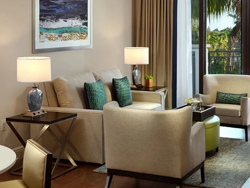 A stylish living room with beige sofas, throw pillows, two lamps, a framed ocean picture, and a balcony view with palm trees.