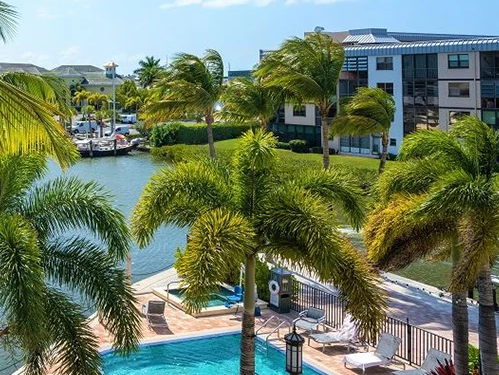 A tropical hotel pool scene with palm trees, a bright blue pool, sun loungers, and a marina nearby, all under a clear sky.
