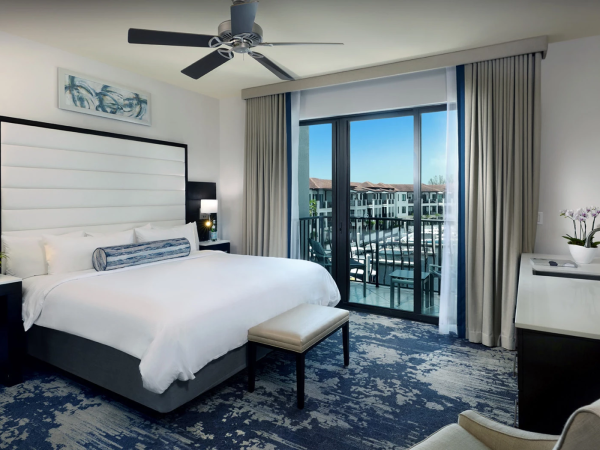 A modern hotel room with a large bed, nightstands, an ottoman, and a balcony overlooking a cityscape, blue patterned carpet, and a ceiling fan.