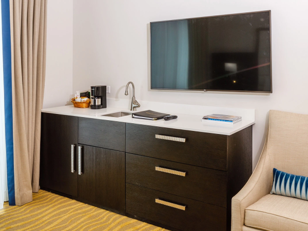 A modern hotel room dresser setup with a wall-mounted TV, coffee machine, kettle, and snacks on a white countertop, plus a dark wood cabinet and a beige chair.
