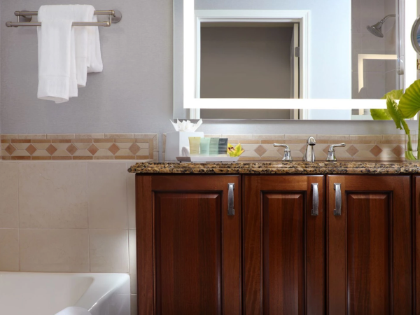 A bathroom scene with a wooden vanity, sink, mirror, towel rack, and a potted plant near the sink, plus a bathtub on the left.