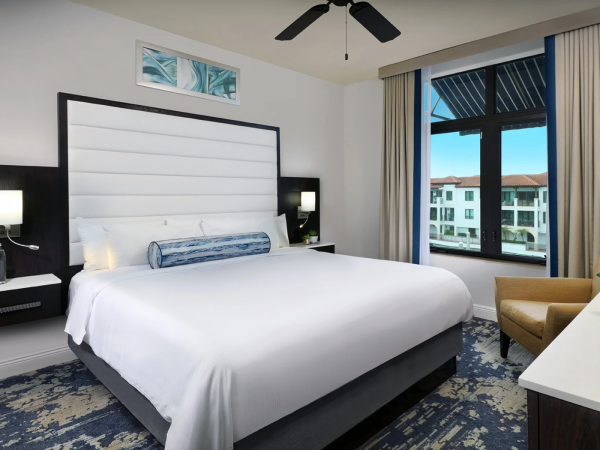 A modern hotel room with a large bed, two nightstands, a white headboard, ceiling fan, window seating, and a blue-carpeted floor.