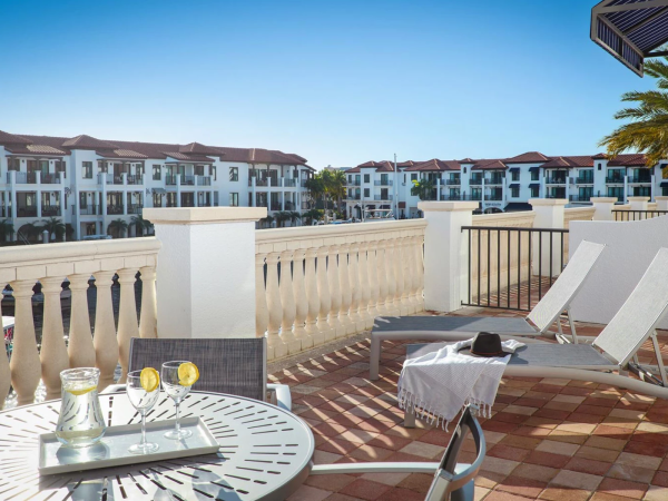 A sunny hotel balcony with two white lounge chairs, a table with drinks, a towel, and a scenic courtyard of white buildings in the distance, bright and relaxing.