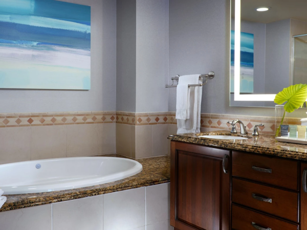 A modern bathroom with a built-in tub, granite countertop, wooden vanity, a large mirror, and a tropical wall art piece, bright and clean.