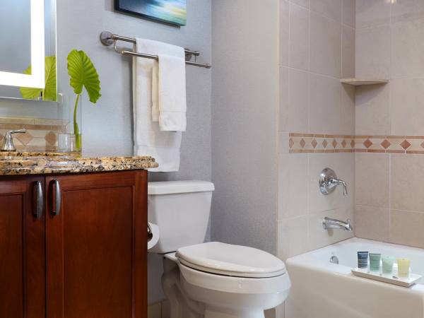 A bathroom with a wooden vanity, granite countertop, towel rack, plant leaf, toilet, and a bathtub with tiled walls and a few toiletries on the edge.