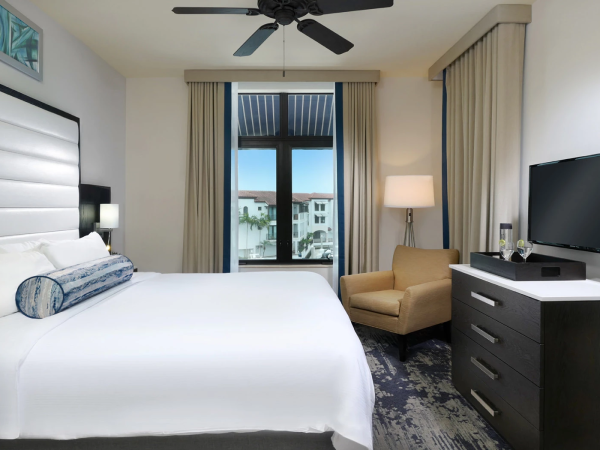 A modern hotel room with a large bed, white linens and pillows, a ceiling fan, a seating chair, a dresser with a flat-screen TV, and a window with view outside.