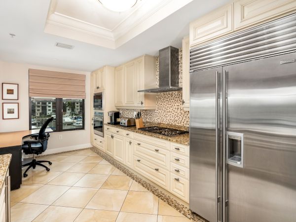 A modern, spacious kitchen with light wood cabinets, granite countertops, tile floor, stainless steel fridge, and a small corner desk by a window, bright and organized.