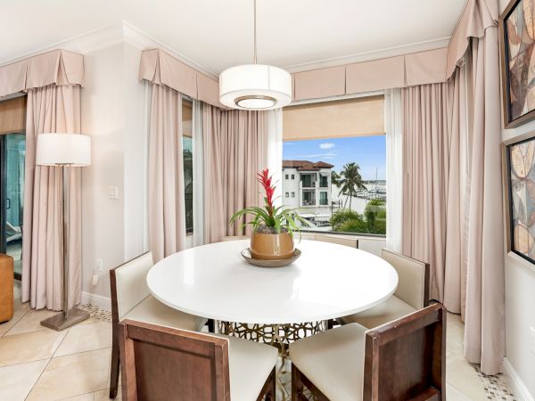 A bright, modern dining nook with a round white table, four cushioned chairs, soft pink curtains, a potted plant centerpiece, and a view of an outdoor pool area through large windows.