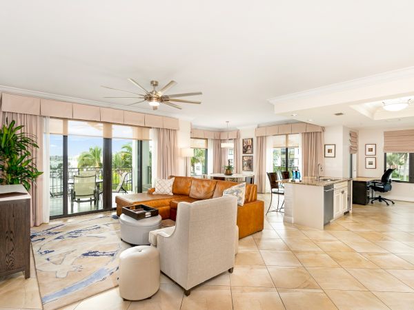Spacious, sunlit open-plan living room with beige tile floors, tan sofa, gray chairs, glass doors to a balcony, and a kitchen area in the background, bright and airy.