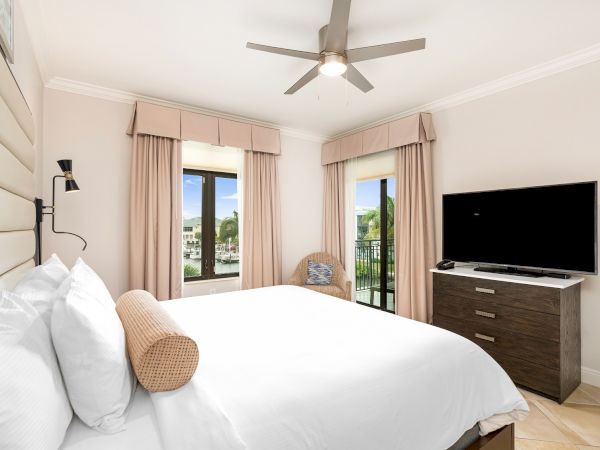 A bright hotel-style bedroom with a large bed, white linens, pink curtains, a ceiling fan, and a dresser with a flat-screen TV.
