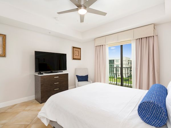 A bright hotel room with a bed, TV on a dresser, window with curtains, a blue chair, ceiling fan, and a balcony view.