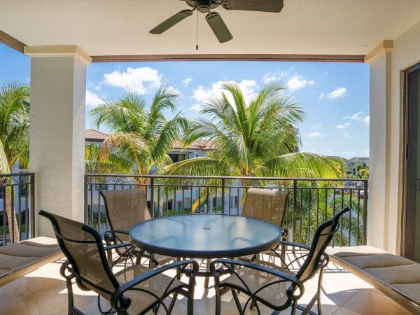 A shaded balcony with a round glass table and metal chairs, overlooking palm trees and a sunny blue sky with a ceiling fan above.