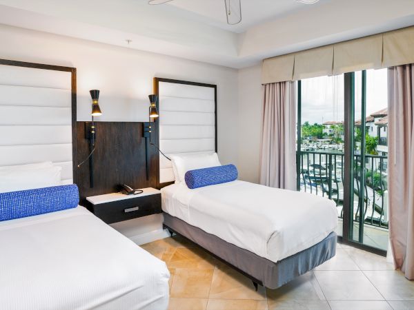 A hotel room with two twin beds, white linens, blue bolster pillows, a shared nightstand, wall lamps, and a balcony with beige curtains.