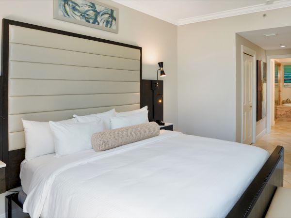 A modern hotel bedroom with a large bed, white linens, two nightstands, ambient wall lamps, and a doorway leading to a bright ensuite.