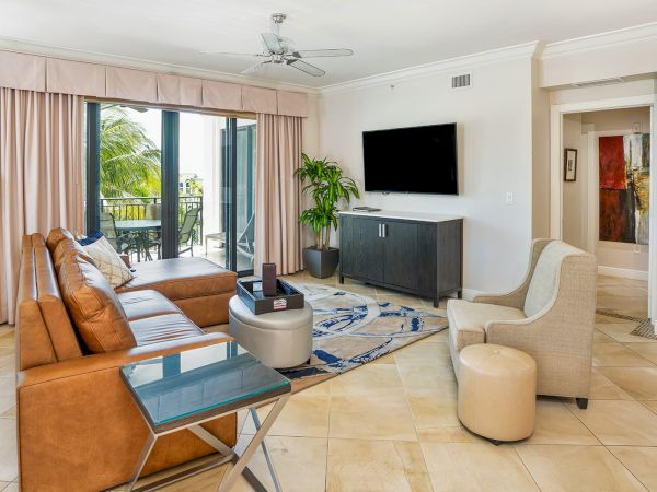 Modern living room with tan leather sofa, light beige armchairs, glass coffee table, TV on a dark console, sliding glass doors to a balcony, and a potted plant.