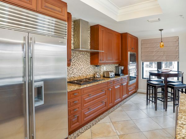 A modern kitchen with stainless steel fridge, wooden cabinets, granite countertops, tiled floor, a small dining nook, and island seating.