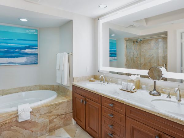 A luxurious bathroom with a marble tub, double vanity, large mirror, and blue ocean artwork on light-toned walls. The space is bright and modern.