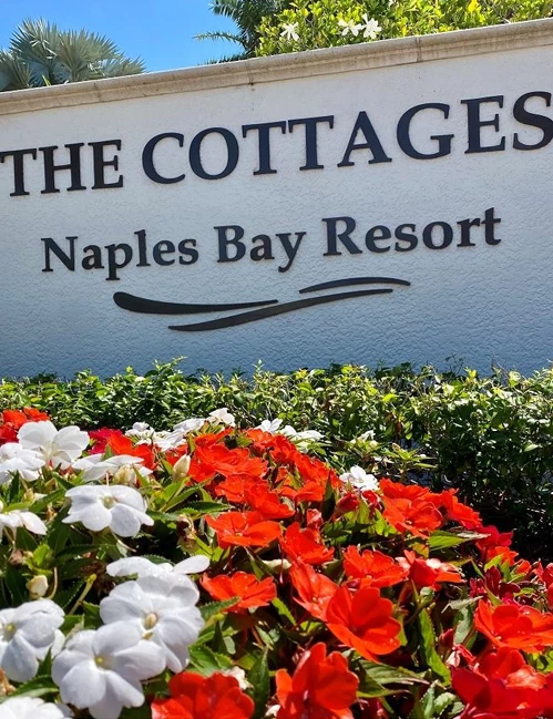 A resort sign reads "The Cottages, Naples Bay Resort" with vibrant red and white flowers in the foreground, tropical shrubbery above.
