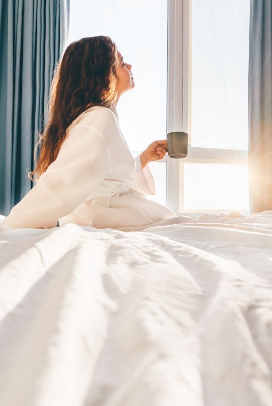 A woman sits up in a sunlit bed by the window, holding a mug and looking out through blue curtains.