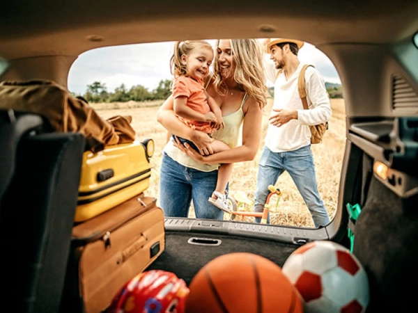 A happy family unloads bright suitcases and toys from a car trunk, with a mother holding a smiling child while another parent stands nearby.