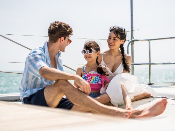 A couple and child relax on a sunny boat, smiling and chatting while the father steers the sail.