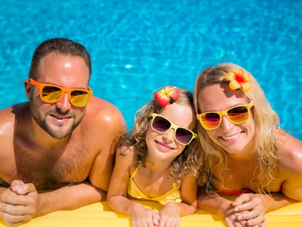 Three people&mdash;two adults and a child&mdash;lean on a pool&rsquo;s edge in yellow swimsuits and sunglasses, smiling under sunny weather.