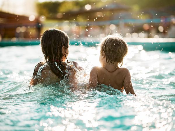 Two kids swim in a sunlit pool, splashing water as they face each other with warm bokeh in the background.