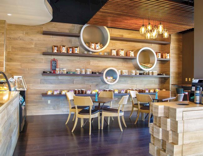A cozy cafe with wooden walls, round wall shelves, hanging lights, and light wood furniture; a small counter on the left and stacked blocks on the right.
