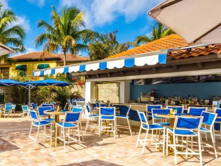 Outdoor tropical bar with blue and white chairs, palm trees, umbrellas, and a tiled patio under a striped canopy, sunny sky.