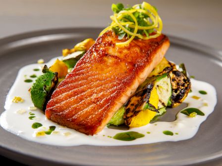 A plated dish features a seared rectangular fish fillet on a creamy white sauce, with greens, charred veg, and citrus garnish topping.