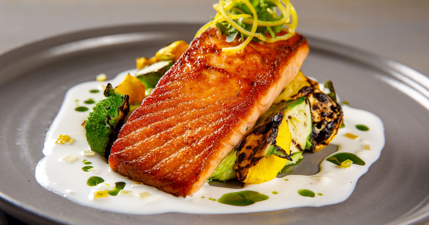 Pan-seared salmon atop grilled vegetables, resting on a creamy white sauce with herbs, citrus zest garnish, and a drizzle of oil.