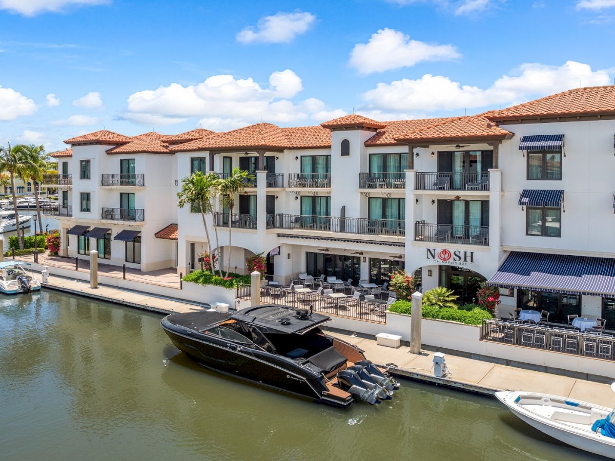 A canalfront shopping and dining row with a white Mediterranean-style building, balconies, palm trees, and a sleek black yacht docked by the sidewalk.