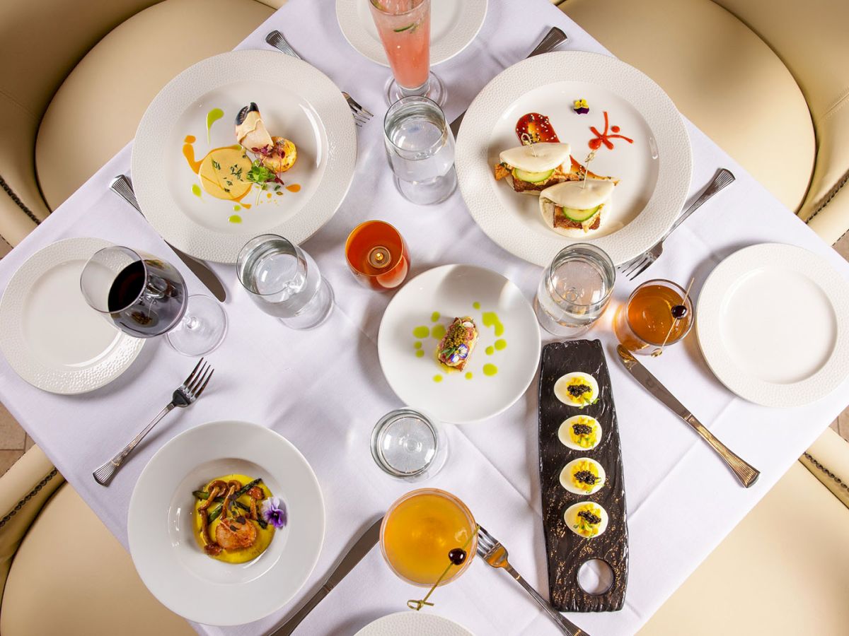 A fancy restaurant table with six plated dishes, drinks, and a long slate tray of bite-sized treats at center, ready for tasting.