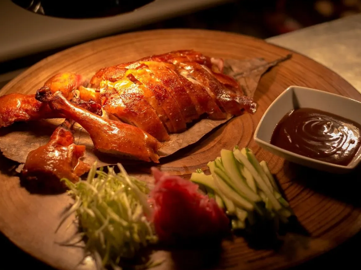 Sliced roasted chicken with crispy skin, shredded cabbage, cucumber sticks, radish, shredded greens, pickled ginger, and a side of brown dipping sauce on a wooden platter, all under warm lighting.