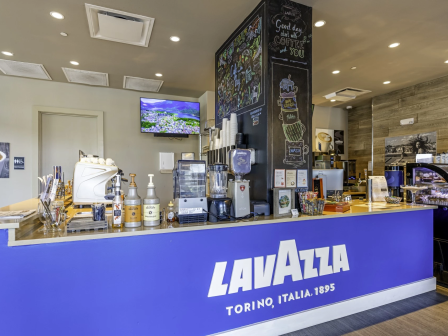 A Lavazza coffee bar setup inside a cafe with a blue counter, espresso machines, syrups, cups, a chalkboard menu, and a TV behind the counter.