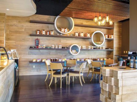 Cozy cafe with wooden walls, circular wall shelves, and a few light fixtures; small tables and chairs sit near a counter on the left.