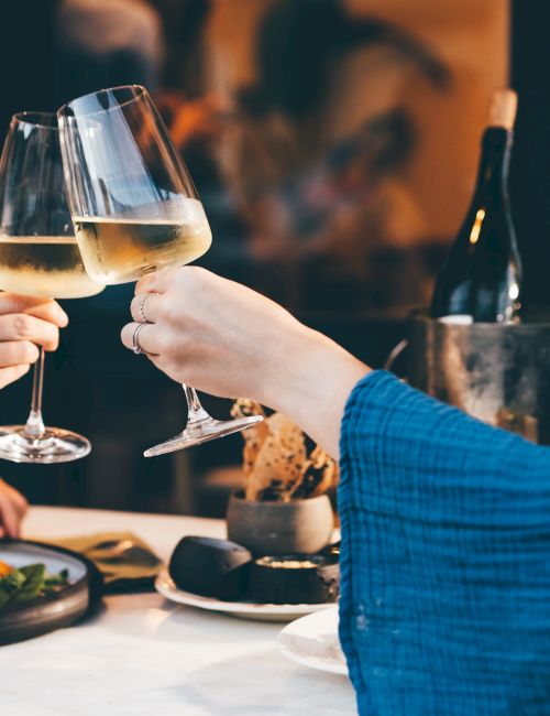 Friends clink two filled wine glasses at a cozy restaurant table, with a bottle and food in the background, celebrating together.