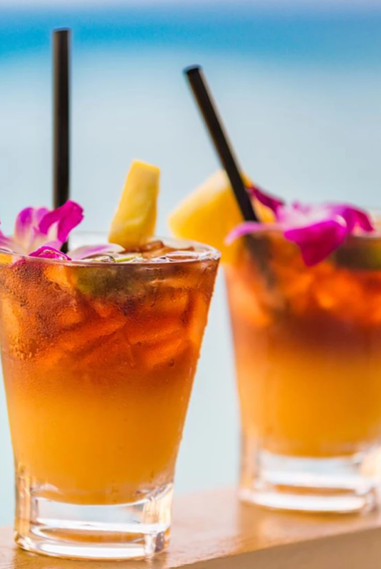 Two tropical cocktails with orange and yellow layers, black straws, and garnish against a sunny beach backdrop.