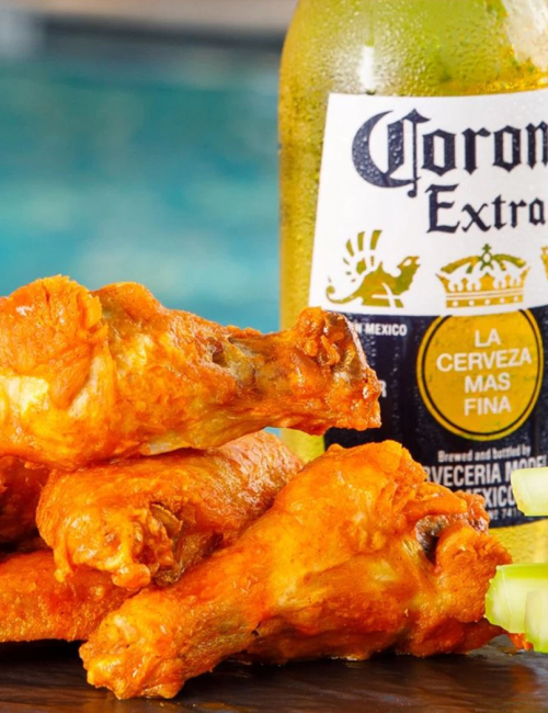 Crispy orange chicken wings on a tray with a Corona Extra bottle beside them, by a poolside backdrop.
