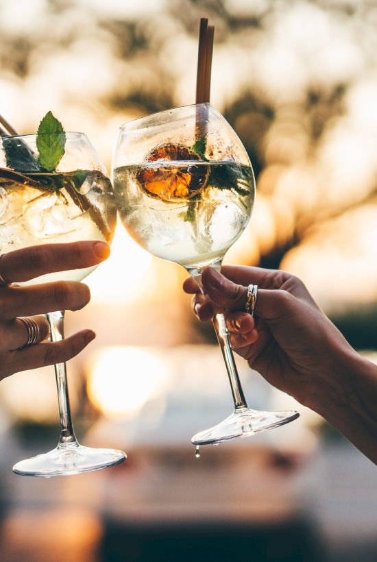 Two people clink glasses of sparkling drinks with mint leaves on a sunny outdoor patio, toasting together in the sunset glow.